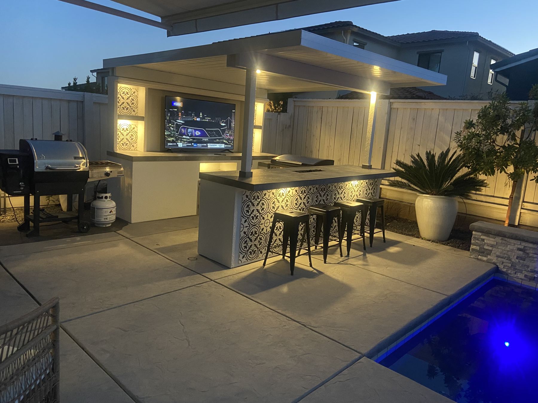 TV island with roof at dusk, lit shelves, under-counter glow on the tiled bar and a lit blue pool