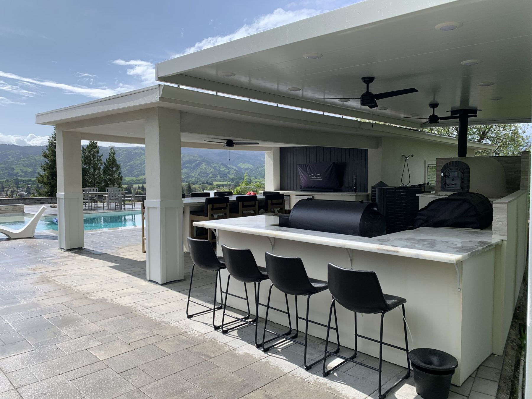 Covered outdoor kitchen with marble bar, four bar stools, smoker, pizza oven and pool with valley views